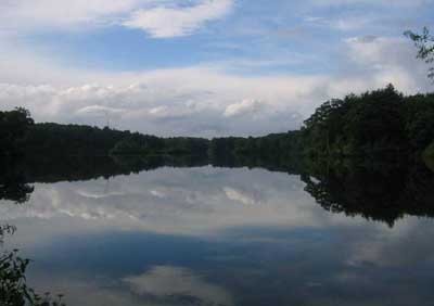 A picture of a mirror-like lake reflecting a blue sky, clouds and the distant shore